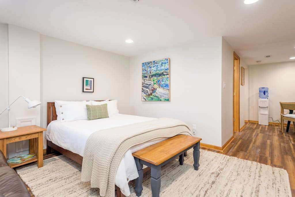 A bedroom with a bed, a bench, a nightstand, a water dispenser, and a painting on the wall.