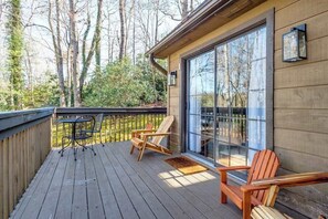 Eine Holzterrasse mit zwei Adirondack-Stühlen und einem Tisch, eine Glastür, die zu einem Raum führt, und Blick auf Bäume.