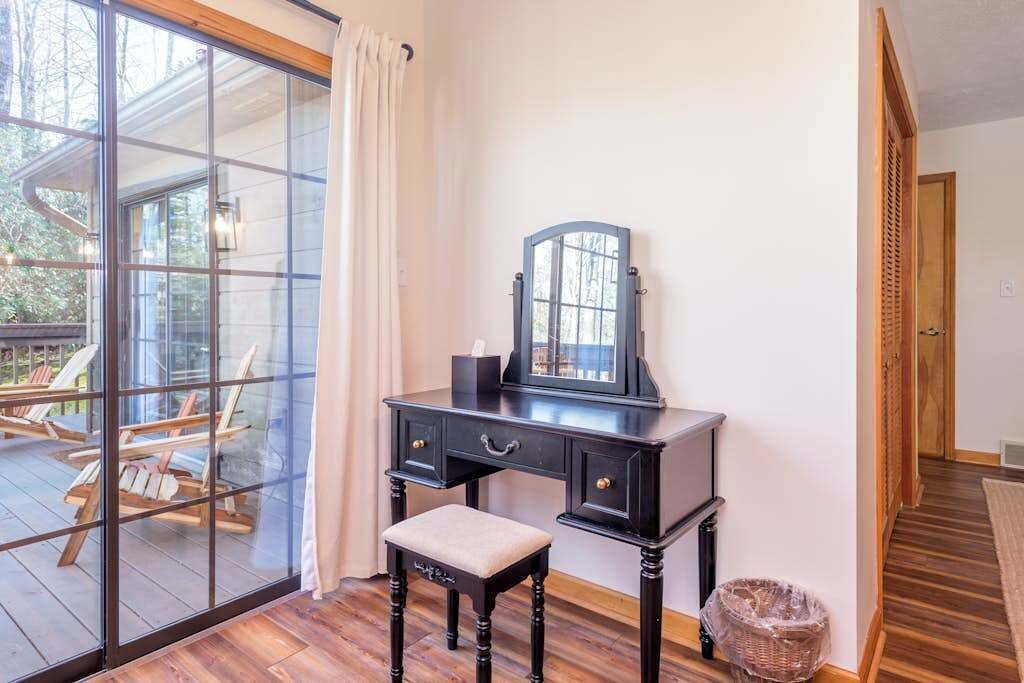 Una habitación con escritorio de madera, una silla, un ventanal con vistas a mesas exteriores, y una puerta que da a otra habitación.