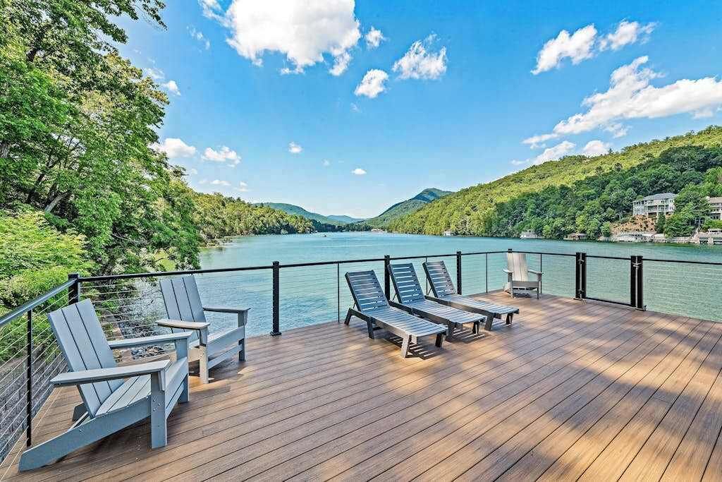 Terraza de madera con sillones y una mesa con vista a un lago, rodeada de follaje y colinas.