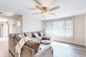 Ein Wohnzimmer mit Ecksofa, rundem Couchtisch, Deckenventilator und einem großen Fenster mit Jalousien.
