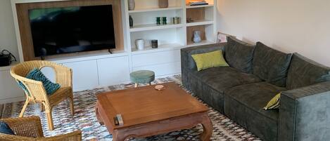 Un salon doté d’un téléviseur à écran plat, de fauteuils en osier, d’un canapé gris, d’une table basse en bois et d’un tapis à motifs.