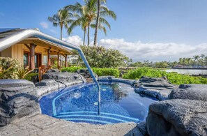 Una piscina con piso de mosaico azul, rodeada de rocas, palmeras y un edificio con techo inclinado.