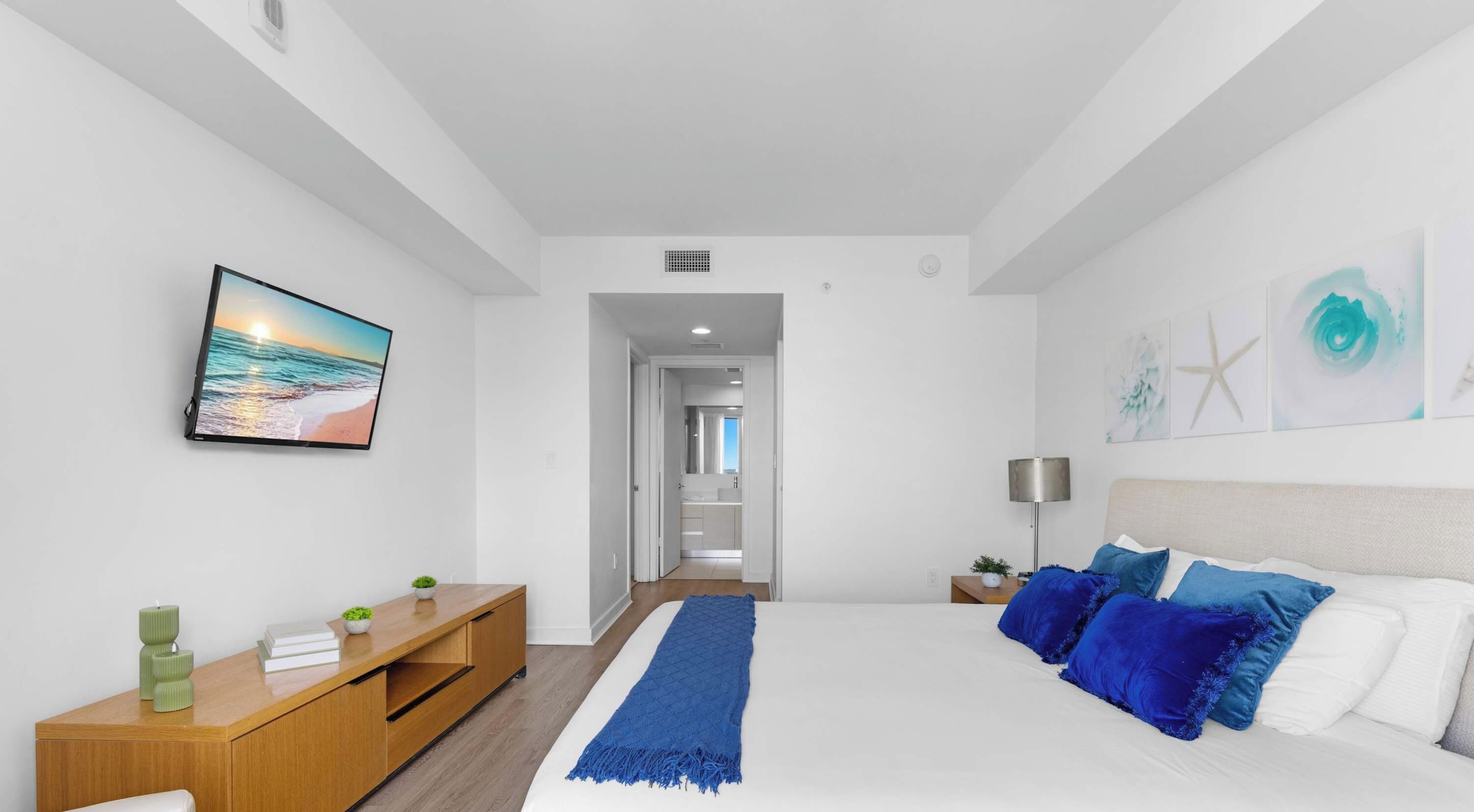 A modern bedroom with a bed, a nightstand, a dresser, a TV, and a hallway leading to a bathroom.