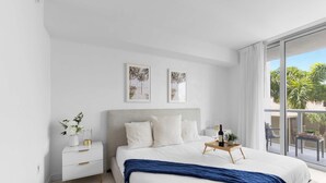 Ein Schlafzimmer mit einem großen Bett, einem Nachttisch, einer Fensterbank und einem Ausblick auf Palmen.