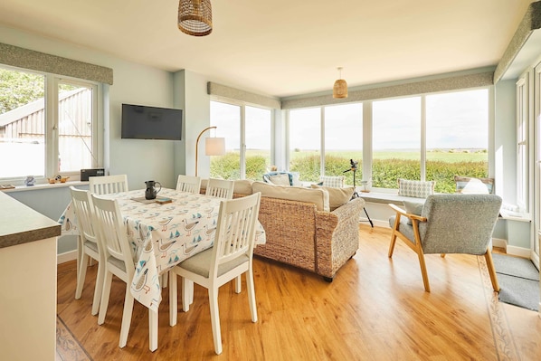 A dining area with a table and chairs, a wicker sofa, and a view through large windows.
