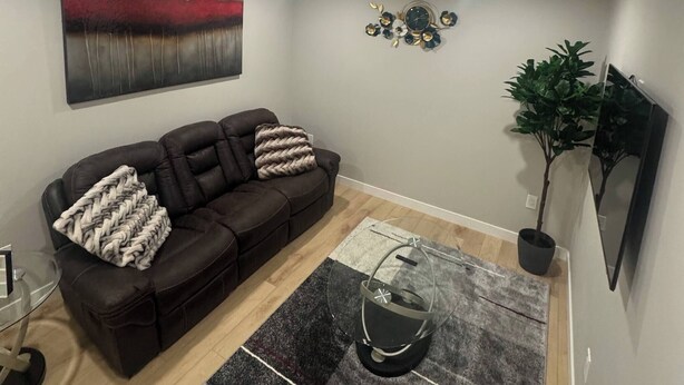 Una sala de estar con un sofá marrón oscuro, una mesa de centro de cristal, un tapete negro y gris, un reloj en la pared y una planta en maceta.