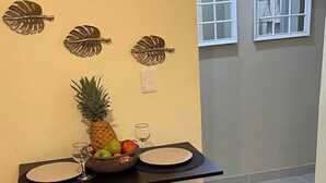 A kitchen with a black countertop, two black stools, a fruit bowl with pineapple and apples, a wine glass, and decorative leaf wall art. - Cozy, Centric Apartment in Central Santo Domingo Este (Santo Domingo Este)