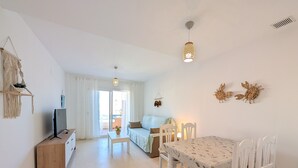 Ein Wohnzimmer mit Fernseher, einer Couch, einem Esstisch mit Stühlen, einer Deckenleuchte und Blick ins Freie.