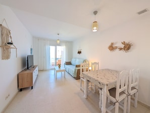 Ein Wohnzimmer mit Fernseher, einer Couch, einem Esstisch mit Stühlen, einer Deckenleuchte und Blick ins Freie.