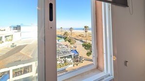 Blick aus einem Fenster auf eine Strandlandschaft mit Palmen, einem Pool und einem Gebäude mit blauem Dach.