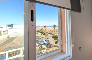 Blick aus einem Fenster auf eine Strandlandschaft mit Palmen, einem Pool und einem Gebäude mit blauem Dach.