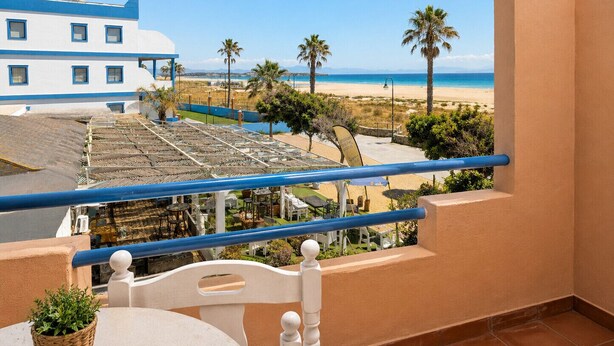 Ein Balkon mit Blick auf einen Strand, Palmen und ein Gebäude mit blau-weißer Fassade.