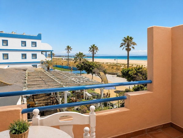 Ein Balkon mit Blick auf einen Strand, Palmen und ein Gebäude mit blau-weißer Fassade.