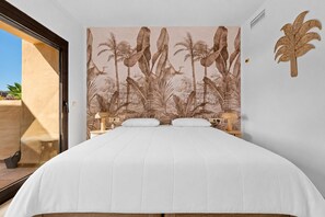 A bedroom with a bed, two bedside tables, a wall mural of tropical foliage, and a door leading to an outdoor area. - Coto Real Penthouse 2470 Naxos Sea View (Manilva)