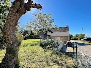 Une maison en pierre avec un toit en pente, entourée d’une cour verdoyante et d’un arbre.