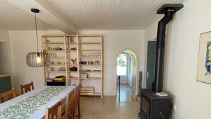 A dining area with a long table, chairs, a hanging light fixture, and a kitchen area with a stove and shelves.