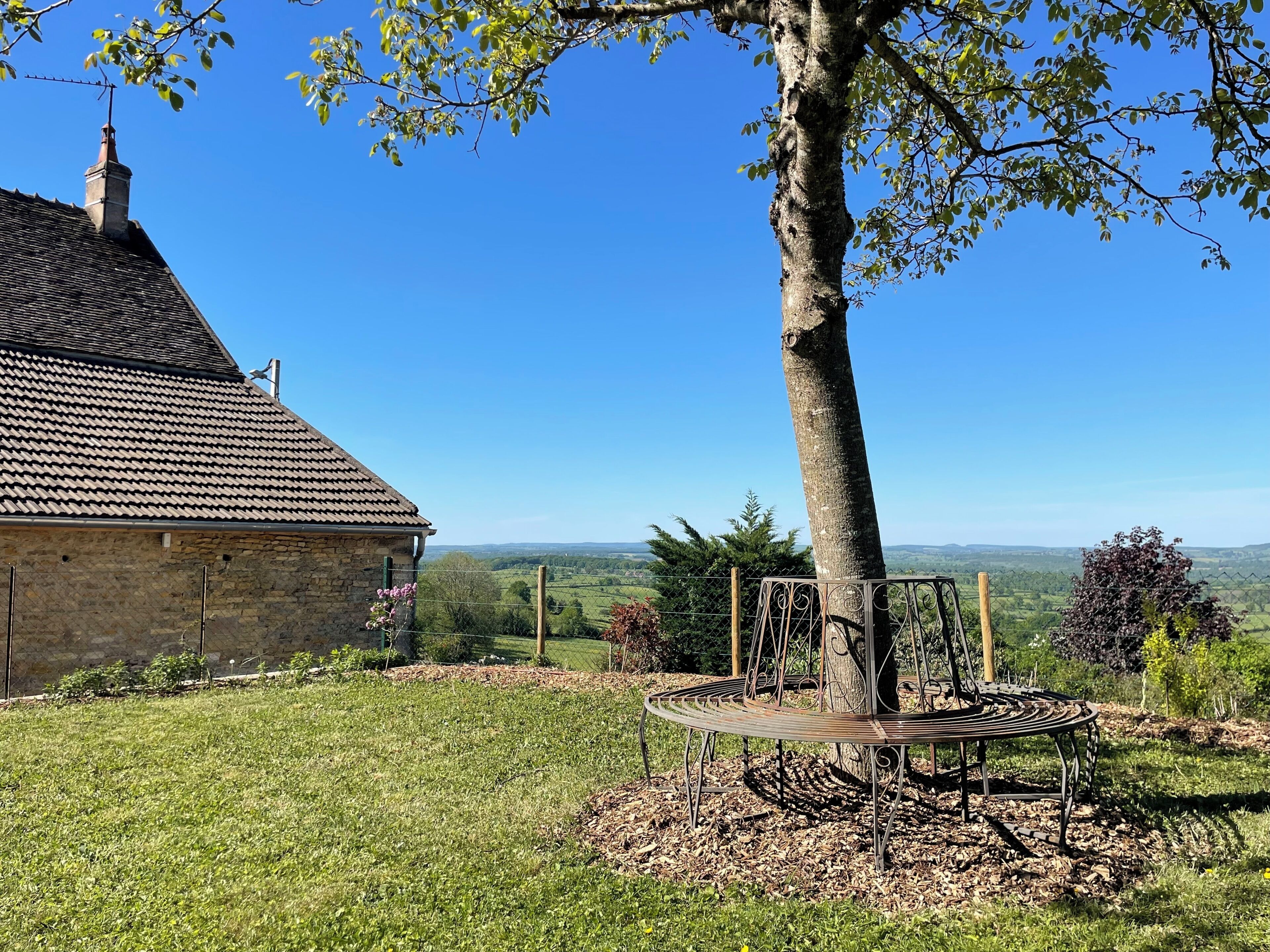 A stone building with a tiled roof, a tree, and a metal bench in a grassy area with a view of distant hills.