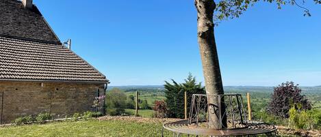 A stone building with a tiled roof, a tree, and a metal bench in a grassy area with a view of distant hills.
