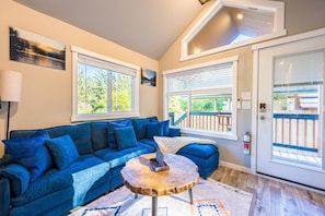A cozy living room with a blue sofa, a round wooden coffee table, and large windows offering views of a deck and greenery. - NEW Luxury Tiny Cabin w/ Hot tub & Fire pit. (Forks)