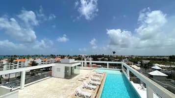 Una piscina en la azotea con sillones de descanso, cielo despejado y vistas de los edificios y palmeras circundantes.