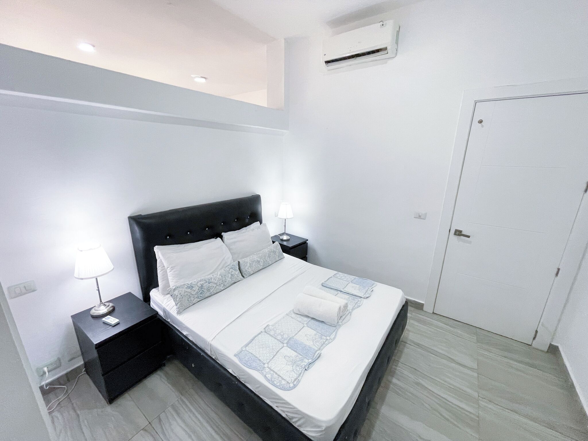 A hotel room with a bed, bedside tables, a nightstand, an air conditioning unit, and a door.