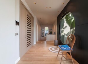 A hallway with a wooden floor, a chair with a patterned cushion, a round rug, and a large window reflecting greenery.