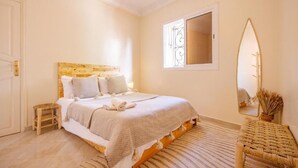 A bedroom with a bed, a bedside table, a mirror, and a window with a view. - Sable Beach Surfcamp Tamraght  (Aourir)