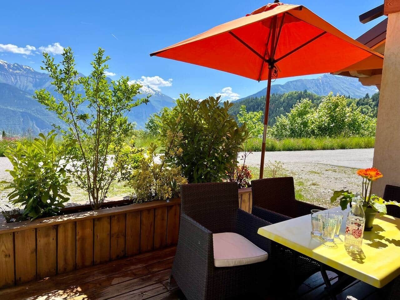 An outdoor seating area with wicker chairs and a table, a red umbrella, and a view of mountains.