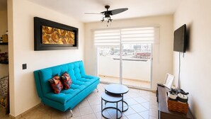 A living room with a blue sofa, a ceiling fan, a TV, and a view through a sliding glass door. - Beautiful apartment with the best location in cabo san lucas 8 min from the beach (Cabo San Lucas)