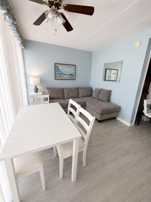 Ein Zimmer mit einem weißen Tisch, weißen Stühlen, einem grauen Sofa, einem Deckenventilator und einem Bild an der Wand.