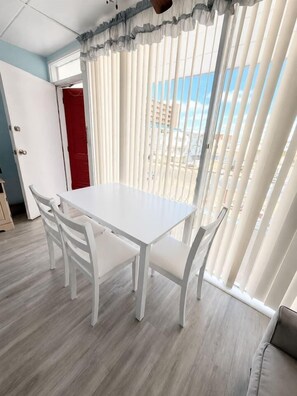 Ein Zimmer mit weißem Tisch und Stühlen, einer roten Tür und Blick durch vertikale Jalousien.