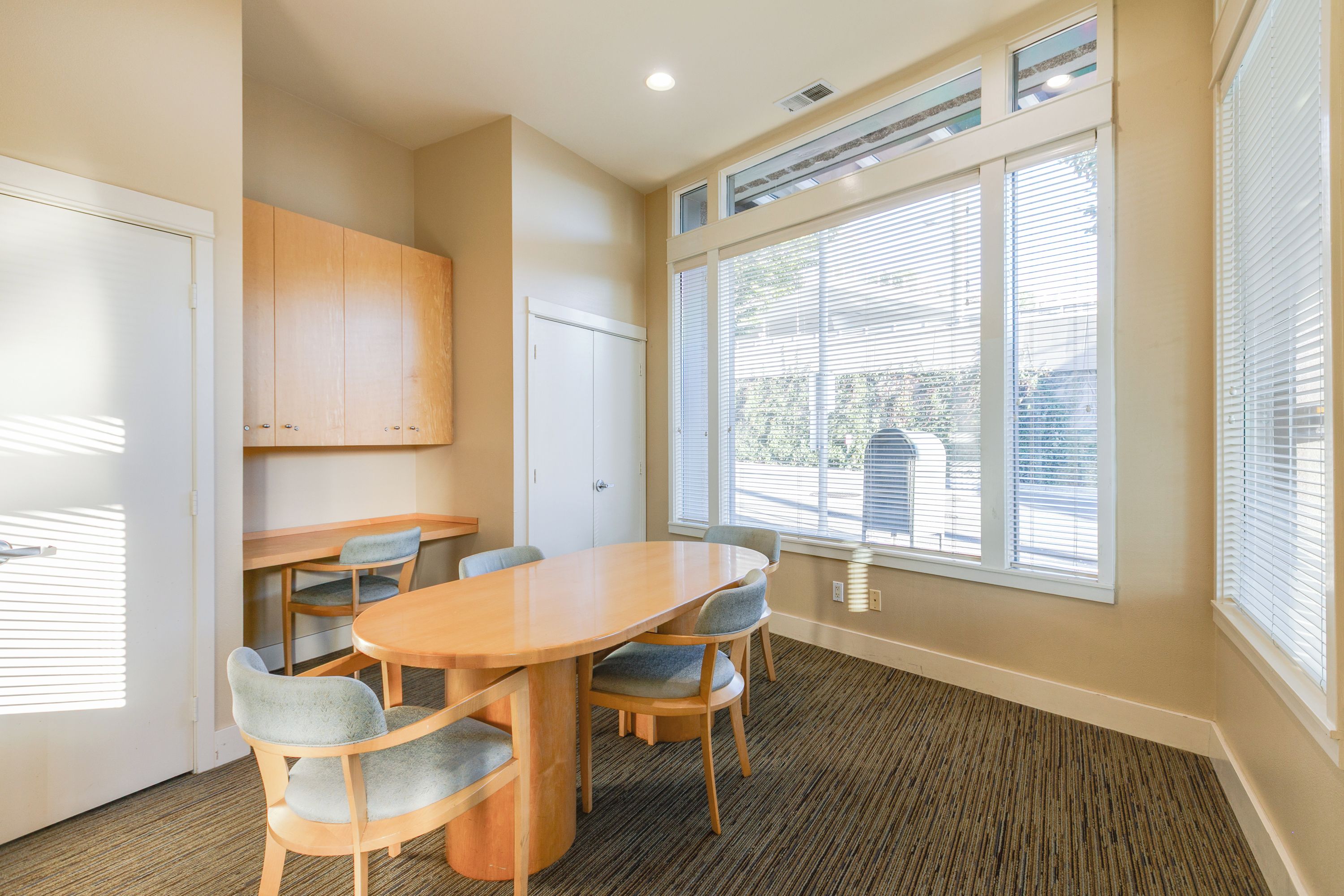 A conference room with a wooden table, four chairs, a white door, and large windows with blinds.