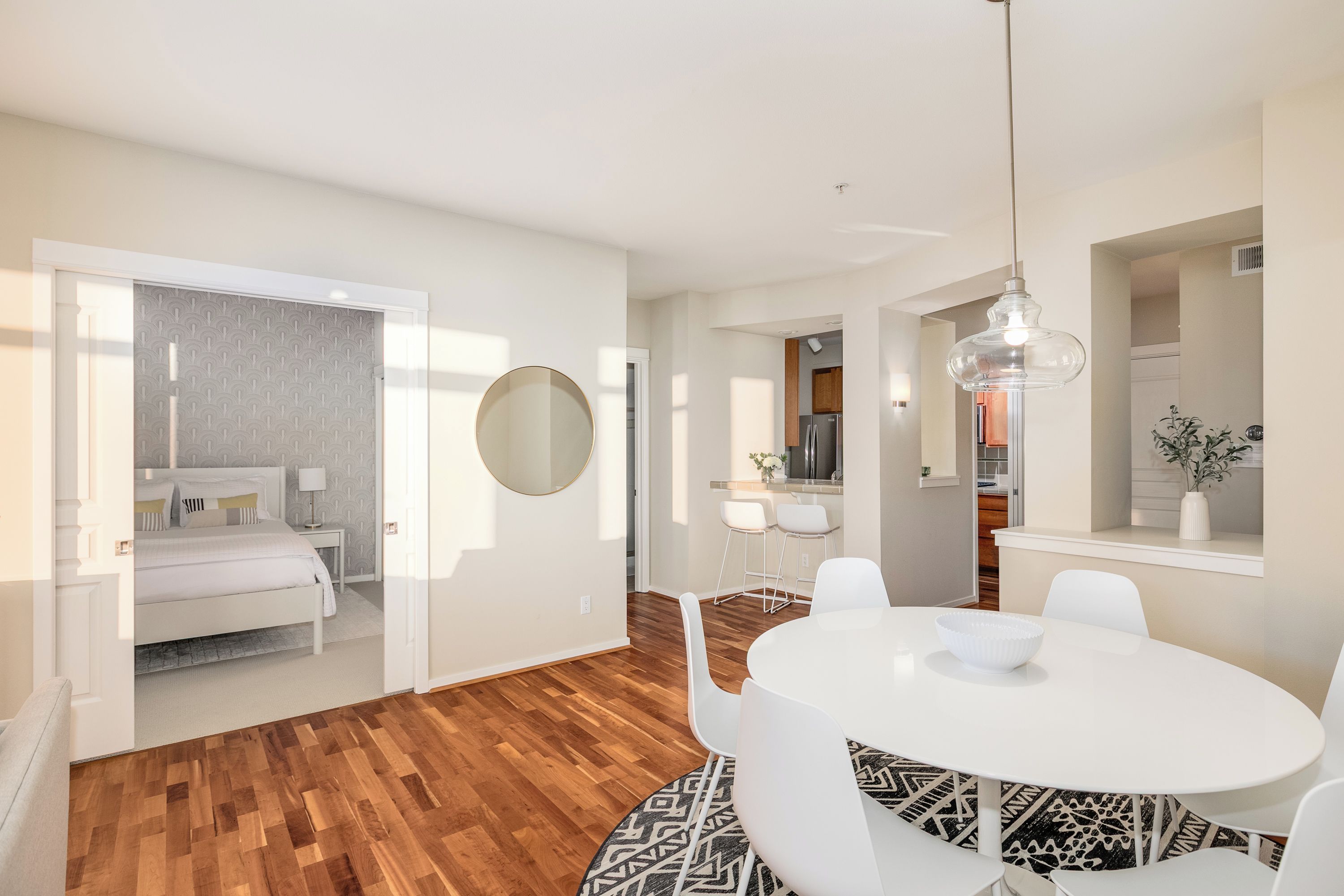 A modern interior with a dining area featuring a round table and white chairs, a kitchen with stainless steel appliances, and a bedroom with a patterned wallpaper and a large mirror.