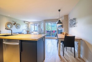 Eine moderne Küche mit einer zentralen Kochinsel, ein Essbereich mit Holztisch und Stühlen sowie Blick auf eine Terrasse mit Gartenmöbeln.
