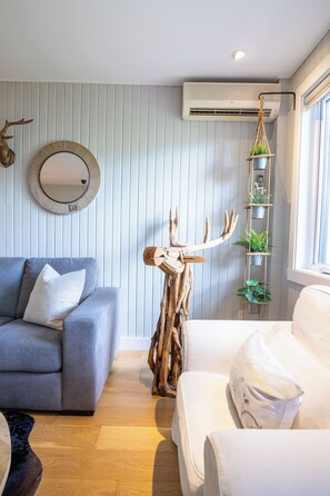 Ein Zimmer mit einem grauen Sofa, einem weißen Sessel, einer Hirschskulptur aus Holz, einem Spiegel und einem Regal mit Pflanzen.