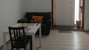 Een eethoek met een tafel, stoelen, een zwarte bank met oranje kussen, een potplant en een lijst met een afbeelding aan de muur.