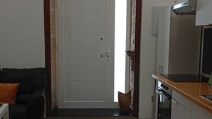 Een ruimtelijk ingerichte leefruimte met een kitchenette, een deur met geëtste glaspanelen, een zwarte bank en een vloerkleed met een patroon.