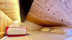 Een kleine, gezellige kamer met een schuine kap, een enkel bed met witte beddengoed, en een rood-wit tas op de vloer.