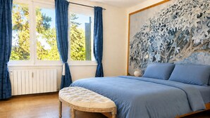 Een slaapkamer met een bed, een raam met gordijnen, een wand met bladmotief en een houten vloer.