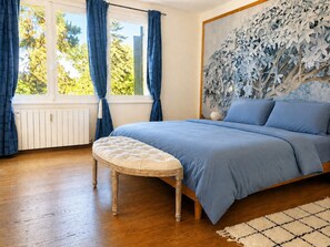 Ein Schlafzimmer mit einem Bett, einem fensterförmigen Ausblick mit Vorhängen, einer Wand mit Blättermotiv und einem Holzboden.