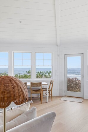 A room with a dining table and chairs, large windows with a view of the sea, and a door leading outside.