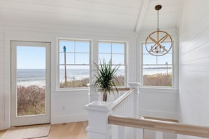 A room with a view of the ocean, white walls, a wooden railing, and a hanging light fixture. - Beachfront Designer Home (Biddeford)