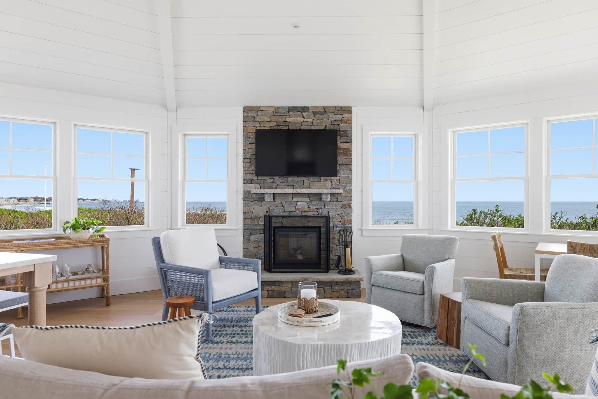 A spacious living room with a stone fireplace, comfortable seating, and large windows overlooking the ocean.