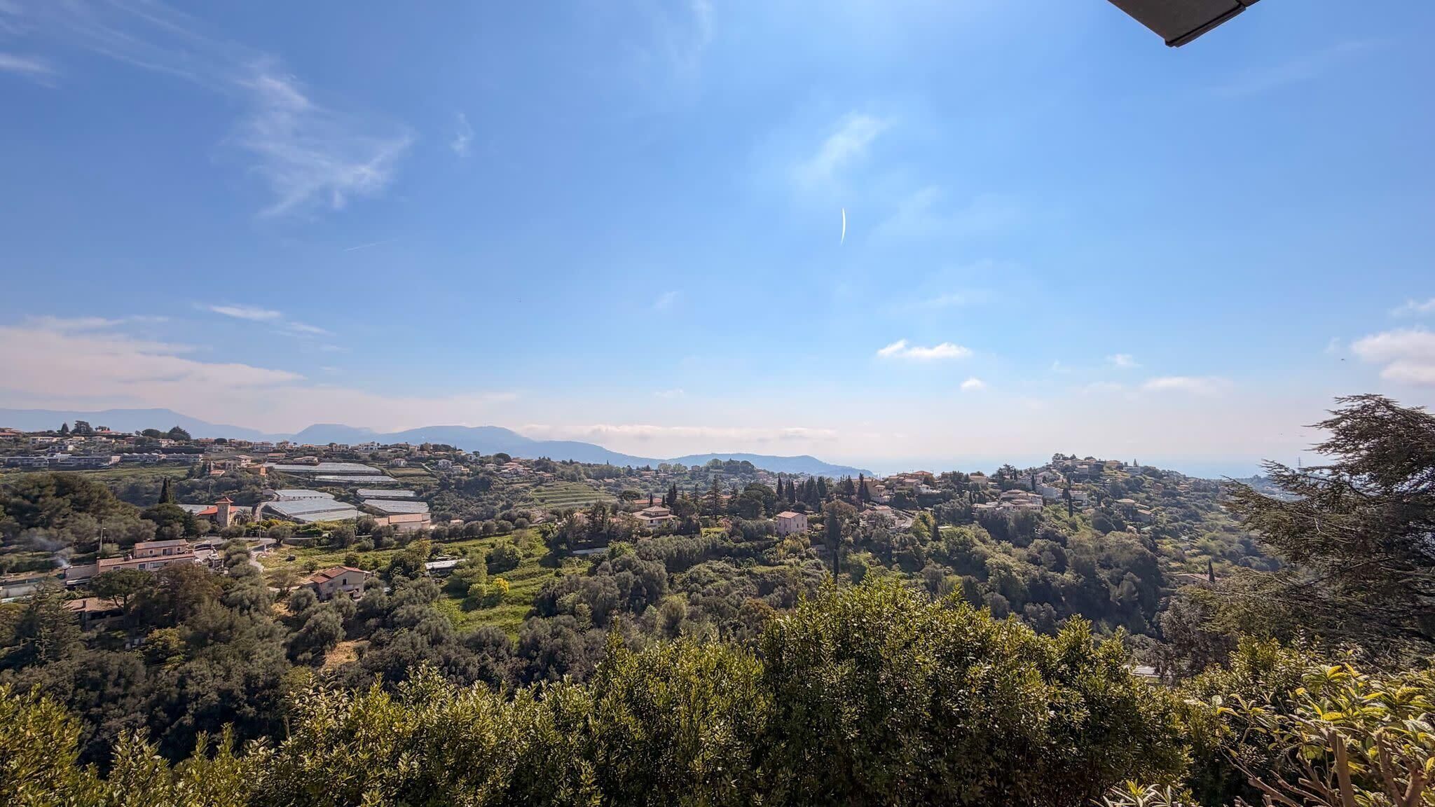 A panoramic view of a hilly landscape with scattered buildings, olive groves, and distant mountains under a clear sky.