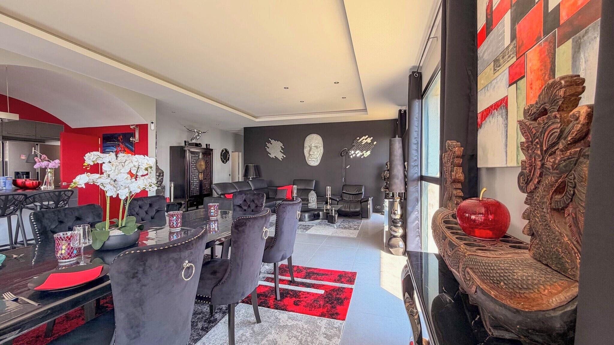A modern living room with a dining area, featuring a red and black color scheme, a large mirror, and decorative wall art.