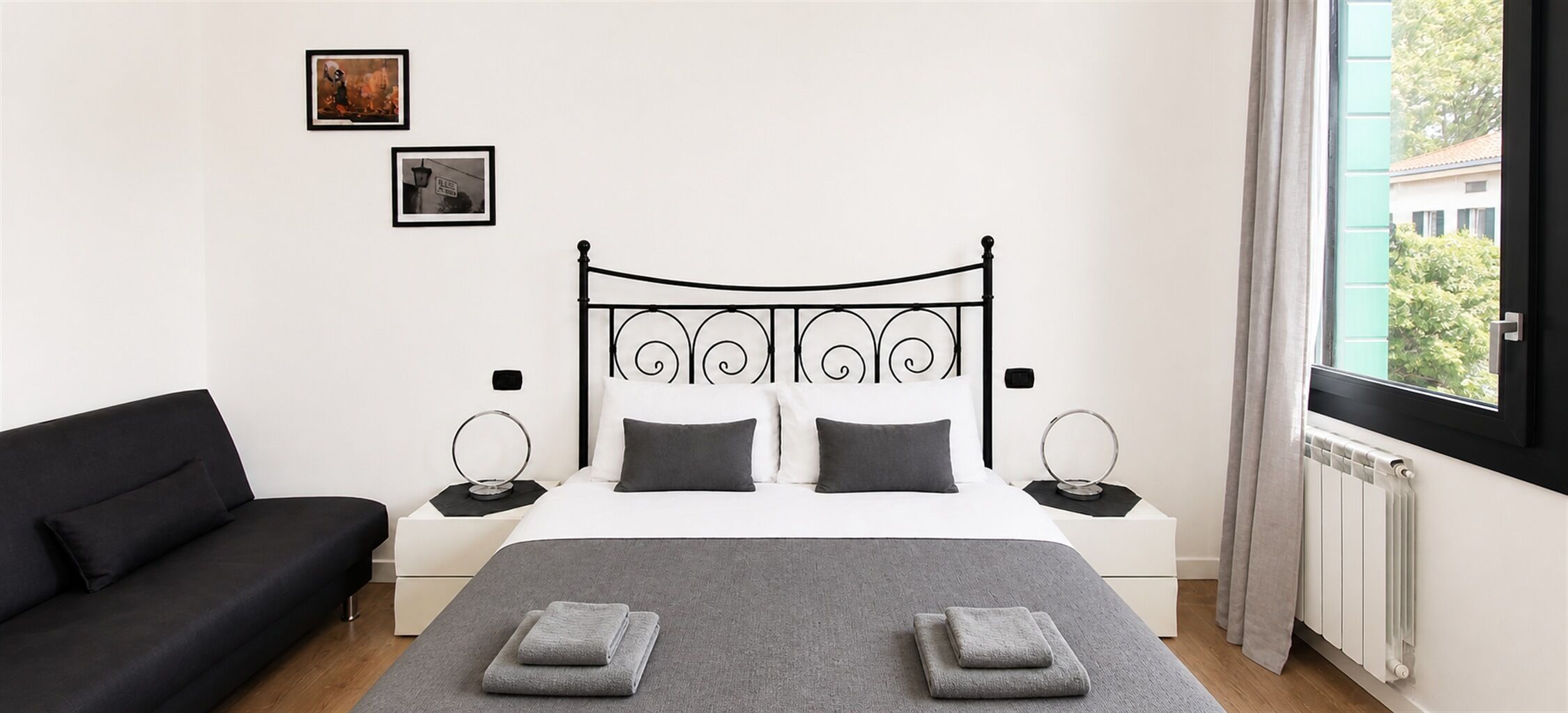 Ein Schlafzimmer mit einem Bett, Nachttischen, einem schwarzen Sofa, einem Fenster mit Vorhängen und zwei gerahmten Bildern an der Wand.