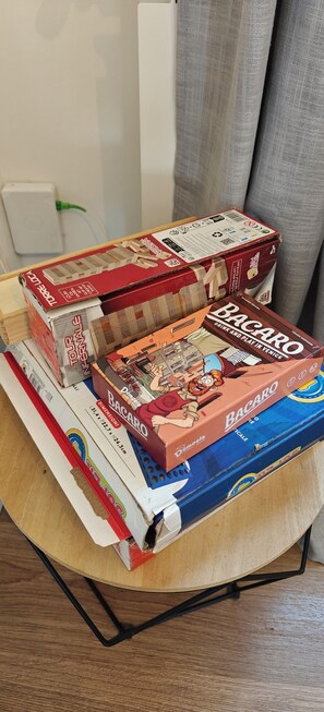 Uma mesa com vários jogos de tabuleiro, incluindo Bacaro, em um quarto com parede branca e cortina cinza.