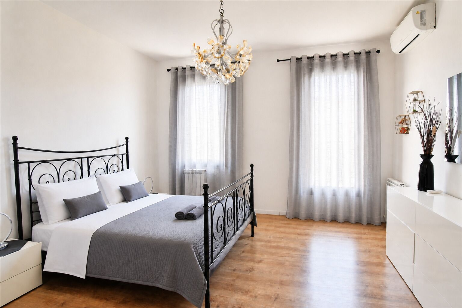 A bedroom with a bed, a chandelier, curtains, a dresser, and an air conditioning unit.