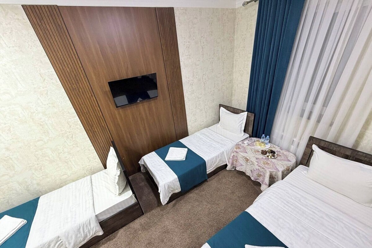 A hotel room with two single beds, a small table with a tablecloth, a TV mounted on a wooden panel, and a window with blue curtains.